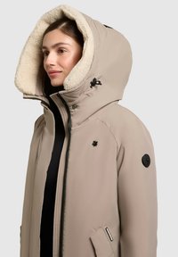 Parka isolata beige con un ampio cappuccio foderato in pelliccia, dotata di pettini neri, tasche con zip e dettaglio logo sul petto e sulla manica.