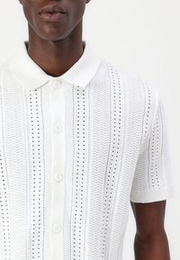 Balta īsām piedurknēm polo kreklā ar izteiktu zigzagu un mazo aciņu rakstu, klasiskā apkaklīte un pogu slēgšana.