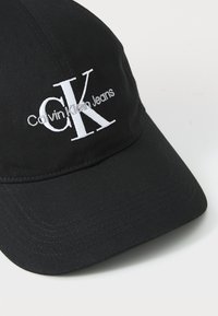 Fekete baseball sapka anyagból, hímzett fehér "Calvin Klein Jeans" logóval és ívelt napellenzővel. Strukturált felület varrási részletekkel.