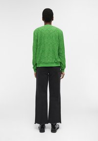 Vihreä neulepusero, jossa on teksturoitu kuvio, yhdistetty mustiin väljään lahkeisiin housuihin ja mustiin kenkiin. Puserossa on ribbireunat hihoissa ja helmassa.