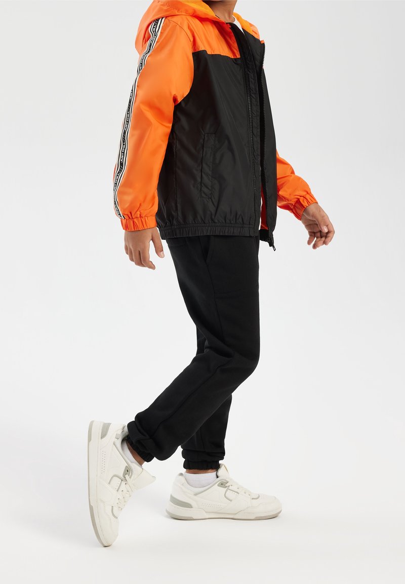 Veste coupe-vent noire et orange avec des poignets élastiques, associée à un pantalon noir et des baskets blanches, comportant des accents gris et un design sportif.