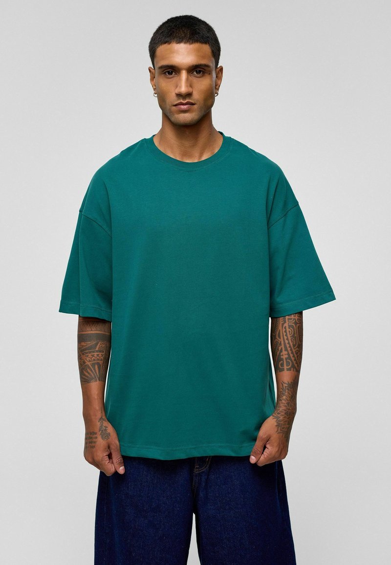 Teal oversized t-shirt met een ronde hals, korte mouwen en een effen textuur. Kenmerkt zichtbare tatoeages op de onderarmen.