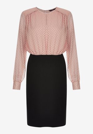 Blouse rose transparente à manches longues avec pois noirs, rentrée dans une jupe noire ajustée jusqu'au genou.