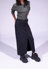 Schwarzer Denim-Maxi-Rock mit einem Schlitz vorne und fransigem Saum, kombiniert mit klobigen schwarzen Sneakern und einem taillierten grauen Rippsweater.