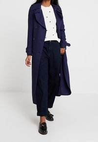 Eine Frau trägt einen langen marineblauen Trenchcoat, einen weißen Pullover mit kleinen schwarzen Punkten, dunkle weit geschnittene Jeans und schwarze Slipper und geht nach vorne.