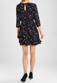 Marinblå klänning med rosa stjärnmönster, trekvartslånga ärmar, nyckelhål bak, bälte i midjan, volangkant, matchad med svarta klackar.