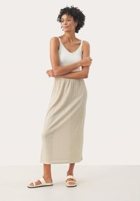 Jupe maxi en beige clair avec une texture ribbée, dotée d'une ceinture élastique. Associée à un débardeur blanc et des sandales plates.