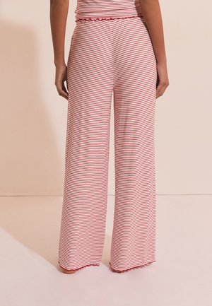 Persona che indossa pantaloni a gamba larga con righe orizzontali rosa e rosse e orli arricciati, in piedi scalza su uno sfondo neutro.