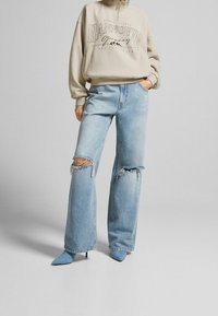 Beige zip-up sweatshirt med texten "HANNOVER", kombinerad med ljusblå slitna vida jeans och blå spetiga klackar.