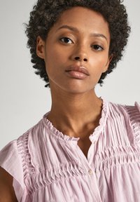 Blouse sans manches rose clair avec un tissu froncé aux épaules et à la poitrine, dotée d'un décolleté arrondi et d'un petit détail de bouton.