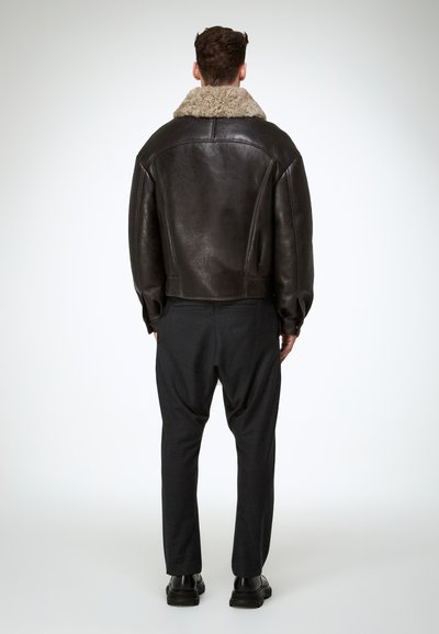 Brown leather jacket with a fluffy beige collar, shown from the back; paired with dark, relaxed-fit trousers and black shoes.