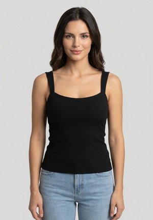 Donna con lunghi capelli scuri che indossa una canottiera nera e jeans azzurri, in piedi davanti a uno sfondo grigio uniforme.