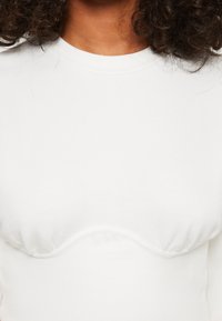 Top bianco a maniche lunghe con scollatura rotonda e texture arricciata sul seno, realizzato in tessuto morbido e elasticizzato.