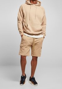 Beige hoodie met een voorzak, gecombineerd met bijpassende cargoshorts met zijzakken. Zwarte en tan sneakers maken de outfit compleet.