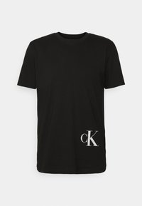 Camiseta de algodón negra con cuello redondo y mangas cortas. Presenta un logo "ck" blanco en la parte inferior. Textura suave, corte estándar.