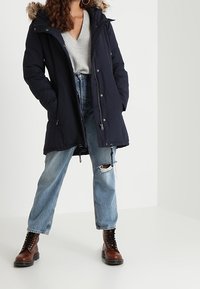 Marinblå parka-jacka med en foder av päls på huvan, med dragkedja och knappstängning, kombinerad med ljusblå jeans med slitningar och bruna snörstövlar.