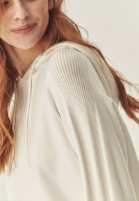 Kremowa hoodie z teksturowanym wykończeniem na ramionach, z kapturem na sznurku i miękkim materiałem. Zbliżenie.