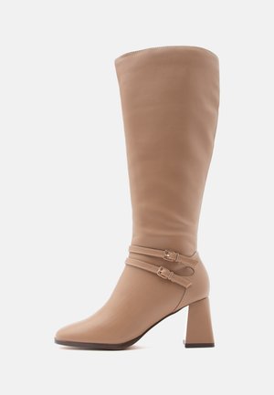 Botte en cuir tan arrivant au genou avec un bout carré, deux sangles décoratives à boucle et un talon large pour plus de stabilité. Texture lisse.