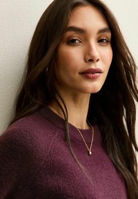 Maglione bordeaux con scollatura rotonda, lunghi capelli scuri e una delicata collana d'oro con un piccolo ciondolo. Carnagione liscia e uniforme.
