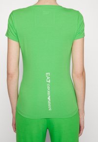 Krótka zielona dopasowana koszulka z bawełny, z okrągłym dekoltem i pionowym białym logo na plecach. Gładka faktura.