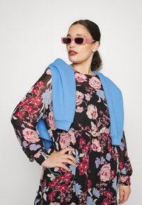 Vestido floral com fundo preto e flores coloridas, combinado com um suéter azul claro pousado sobre os ombros e óculos de sol cor-de-rosa.