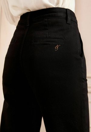Pantalons noirs avec une texture lisse, arborant un petit logo brodé en cuivre. Comprend deux poches arrière et un design de ceinture classique.