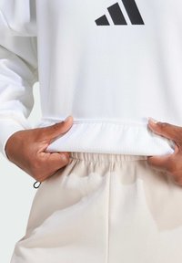 Biele mikina s čiernym logom, zdobená textúrovaným lemom. Skombinovaná so svetlými béžovými nohavicami s elastickým pásom, ktoré majú uvoľnený strih.