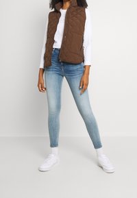 Brun kviltad väst över en vit långärmad tröja, tillsammans med ljusblå skinny jeans och vita sneakers. Enkelt design, inga synliga mönster.