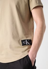 Tricou din bumbac bej cu mâneci scurte, cu un eticheta neagră cu logo alb și o croială lejeră. Materialul pare neted �și casual.