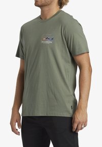 Olive green short-sleeve t-shirt made of cotton. Features a rectangular graphic with mountains and "BILLABONG" text in blue and yellow.