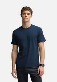 T-shirt blu navy a maniche corte realizzato in tessuto liscio con scollatura rotonda e cuciture minime, abbinato a jeans scuri e un orologio nero.