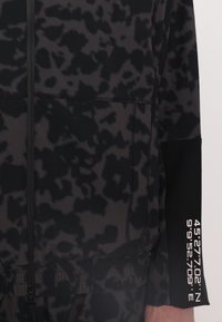 Black jacket featuring a textured pattern, zip closure, elastic hem, and printed coordinates on the sleeve. Smooth fabric with subtle sheen.