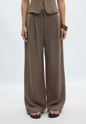 Personne portant un pantalon large marron texturé, un gilet sans manches assorti, des sandales et des bracelets marron à un poignet.