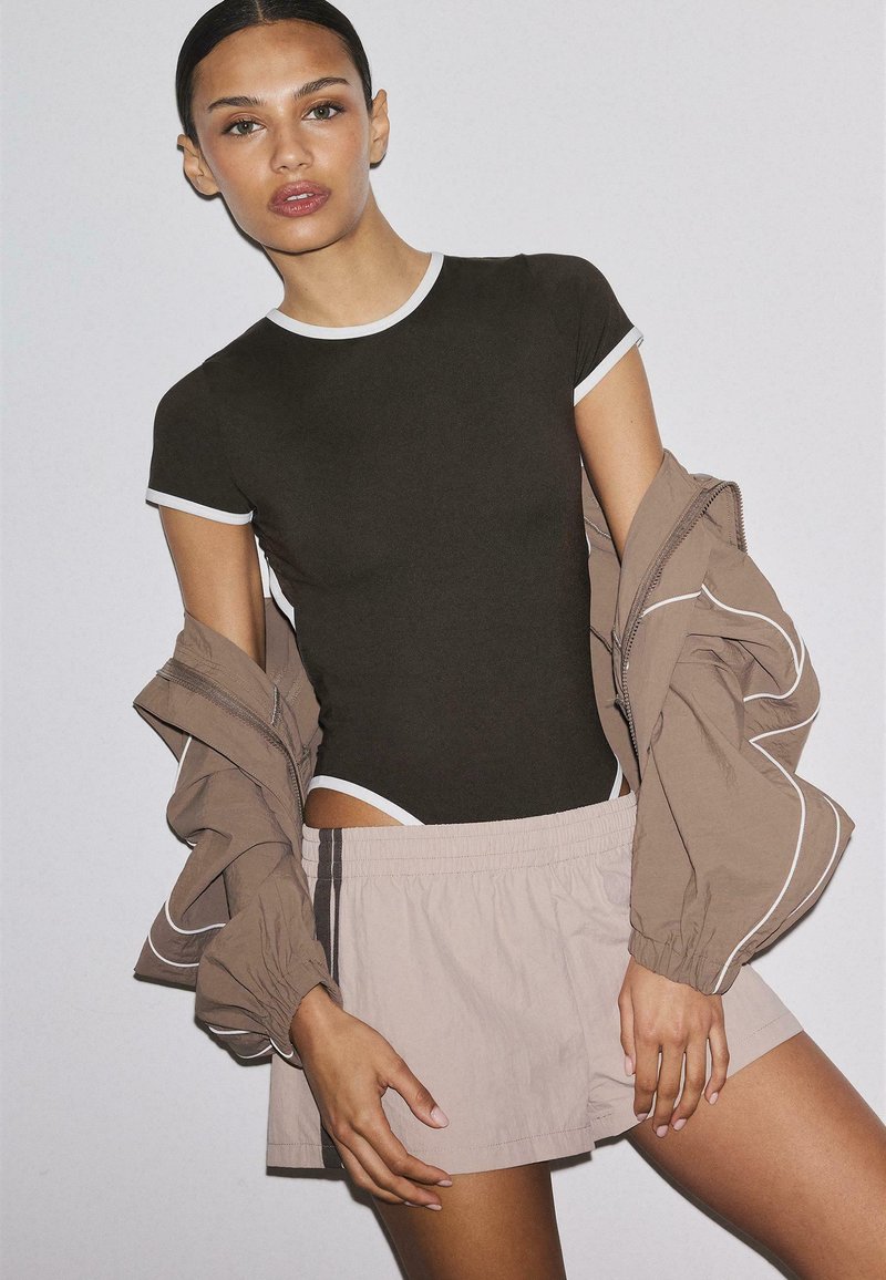Femme portant une chemise noire ajustée avec des bordures blanches, un short beige et une veste marron partiellement portée avec des liserés blancs, debout devant un fond uni.