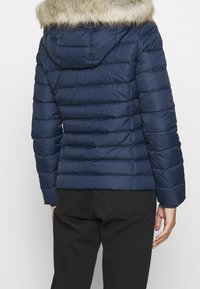 Person in einer marineblauen gesteppte Jacke mit kapuze aus fellbesatz und schwarzen Hosen, von hinten vor einem schlichten Hintergrund gezeigt.