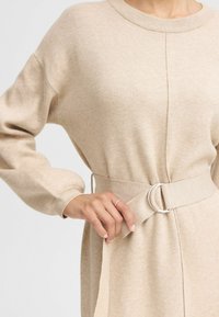 Beiges Strickkleid mit rundem Ausschnitt, langen Ballonärmeln und einem Stoffgürtel mit silbernem Ringakzent. Weiche Textur und lockere Passform.