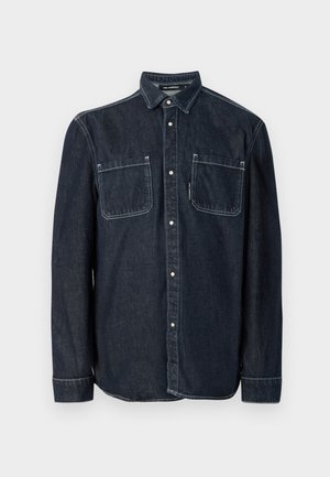 Camisa de mezclilla oscura de botones con cuello puntiagudo, dos bolsillos en el pecho y acentos de costura blanca. Mangas largas con puños abotonados.