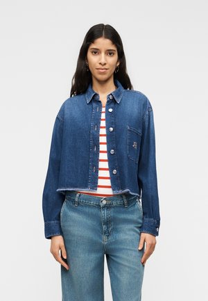 Naine, kes kannab lühikest sinist teksajakki punase-valge triibulise särgi ja hele siniste teksade üle, seisab ühevärvilise tausta ees.