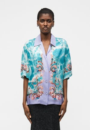 Frau trägt ein kurzärmeliges Hemd mit Knopfleiste mit blau-lila barocken und wasserinspirierten Mustern, kombiniert mit einem dunklen Rock.