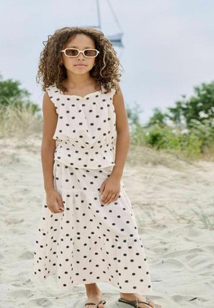 Ung jente iført hvit kjole med svarte prikker og solbriller står barfot på en sandstrand med grønt i bakgrunnen.