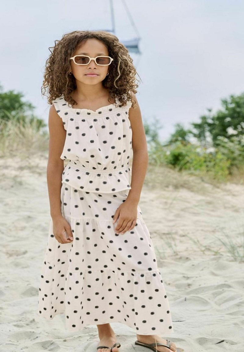 Jeune fille portant une robe blanche à pois noirs et des lunettes de soleil, debout pieds nus sur une plage de sable avec de la verdure en arrière-plan.