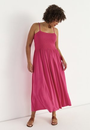 Frau mit lockigem Haar, die ein ärmelloses, bodenlanges pinkfarbenes Kleid und braune flache Sandalen trägt, steht vor einem weißen Hintergrund.