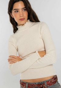 Jersey de cuello alto en color crema con canalé y cuello y puños con volantes. Diseño ajustado con mangas largas. Combinado con un cinturón estampado sobre pantalones grises.