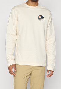 Pitkähihainen kerma-valkoinen paita pehmeästä kankaasta, pyöreällä kaula-aukolla. Rintataskussa pieni musta ja persikkainen Patagonia-logo.