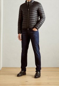 Vadderad svart jacka med horisontell söm, dragkedja fram, tillsammans med mörkblå jeans och bruna skor med spänne.