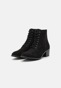Botas de tornozelo em camurça preta com biqueira arredondada e salto bloco. Apresentam um fecho de atacadores na frente e ilhós de metal. Superfície texturizada sem padrões.