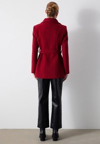 Abrigo rojo de mezcla de lana con cintura ceñida, hombros estructurados, costuras en la parte posterior, usado sobre pantalones de cuero acolchados negros y tacones.