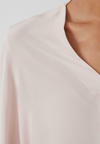 Blouse rose clair avec une texture lisse, dotée d'un décolleté en V peu profond et d'un tissu drapant en douceur. Les bords sont soigneusement finis.