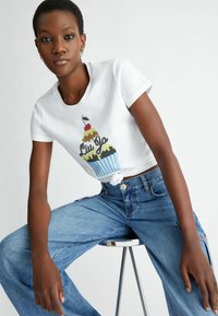 Camiseta blanca de manga corta con un gráfico de cupcake con detalles multicolores y un frente anudado. Jeans de mezclilla anchos de color azul.