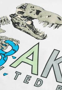 Weißer Stoff mit einem Druck eines Dinosaurierschädels in Grau und Schwarz, mit stilisiertem Text "BAK" in Blau und Grün sowie dekorativen Akzenten.